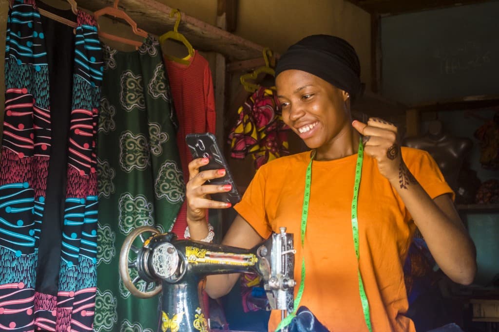 Nigerian fashion entrepreneur at her sewing machine, smiling at her phone after a BizFunding approval.